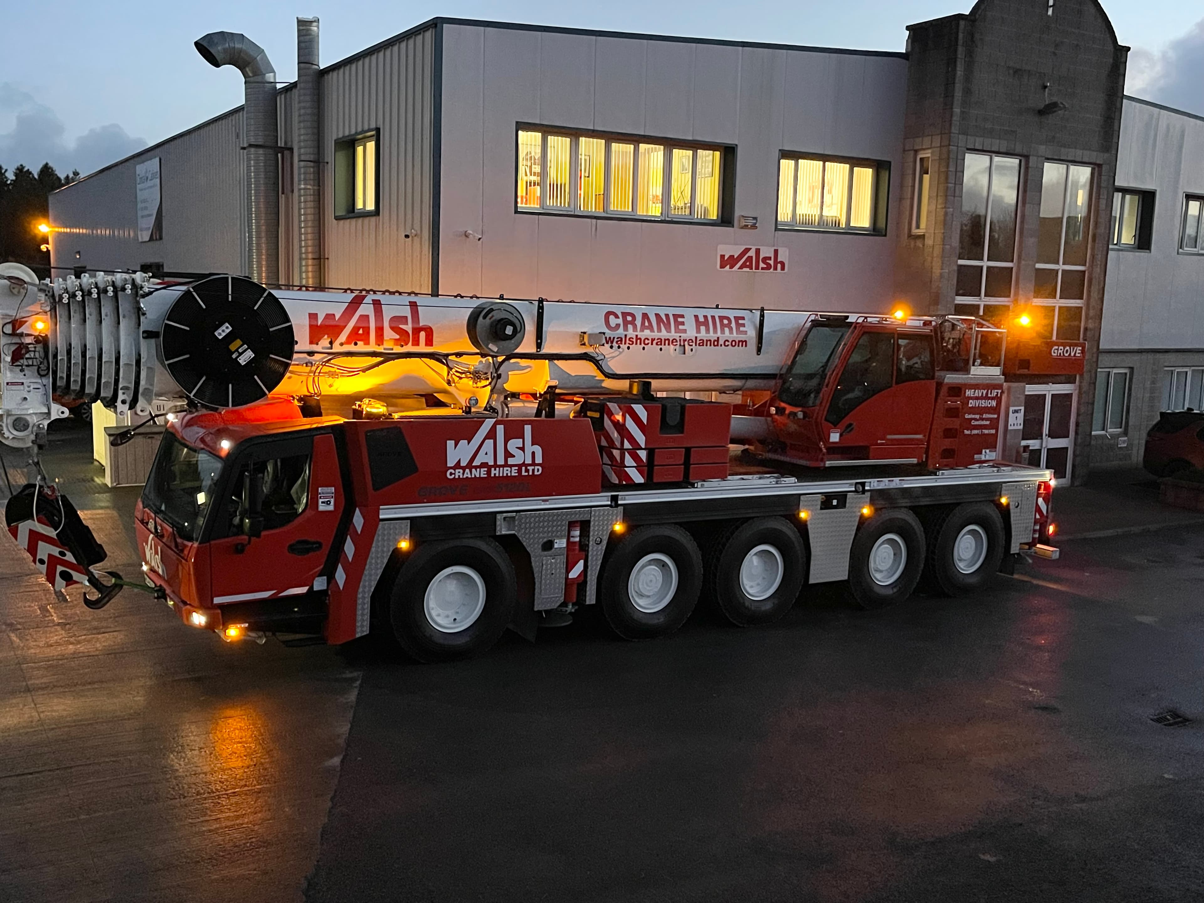 large multi-axle mobile crane truck with "Walsh Crane Hire" branding, parked outside an industrial building at dusk