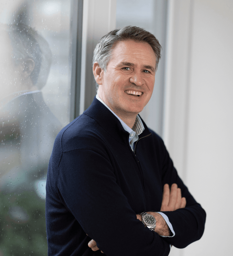 Man with grey hair in navy blazer, smiling, indoors near a window