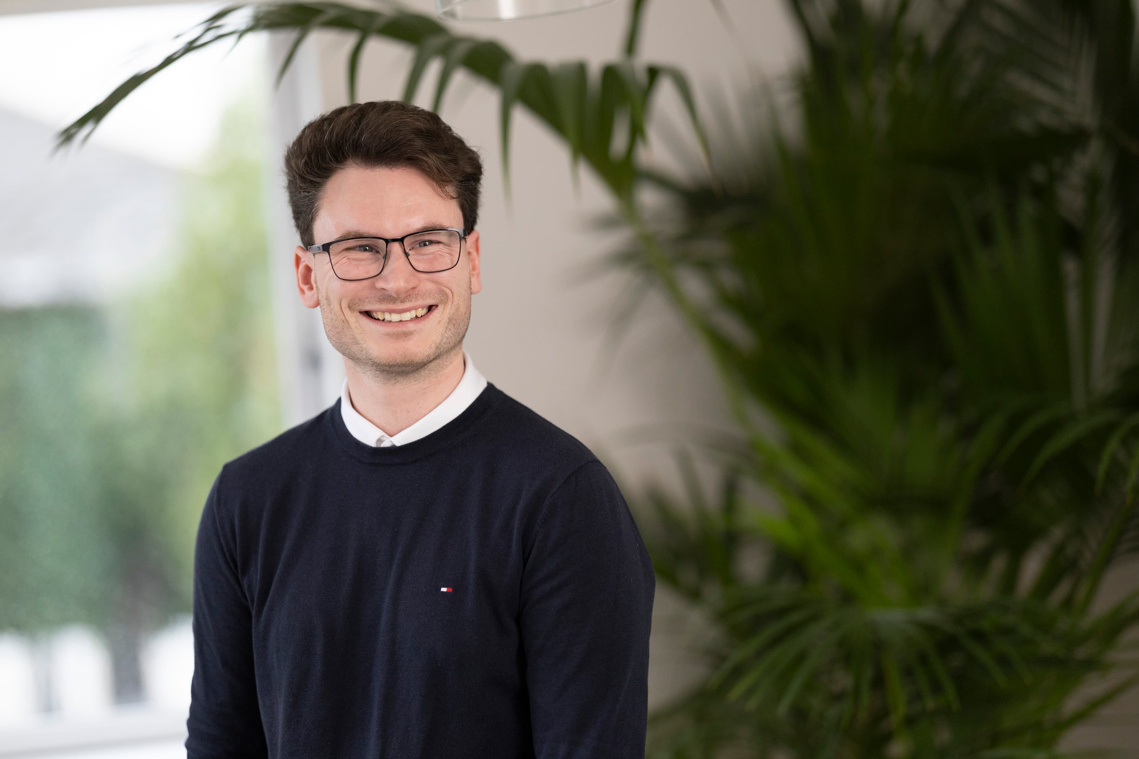 Young man with glasses and light beard, smiling, dark jumper, plant in background