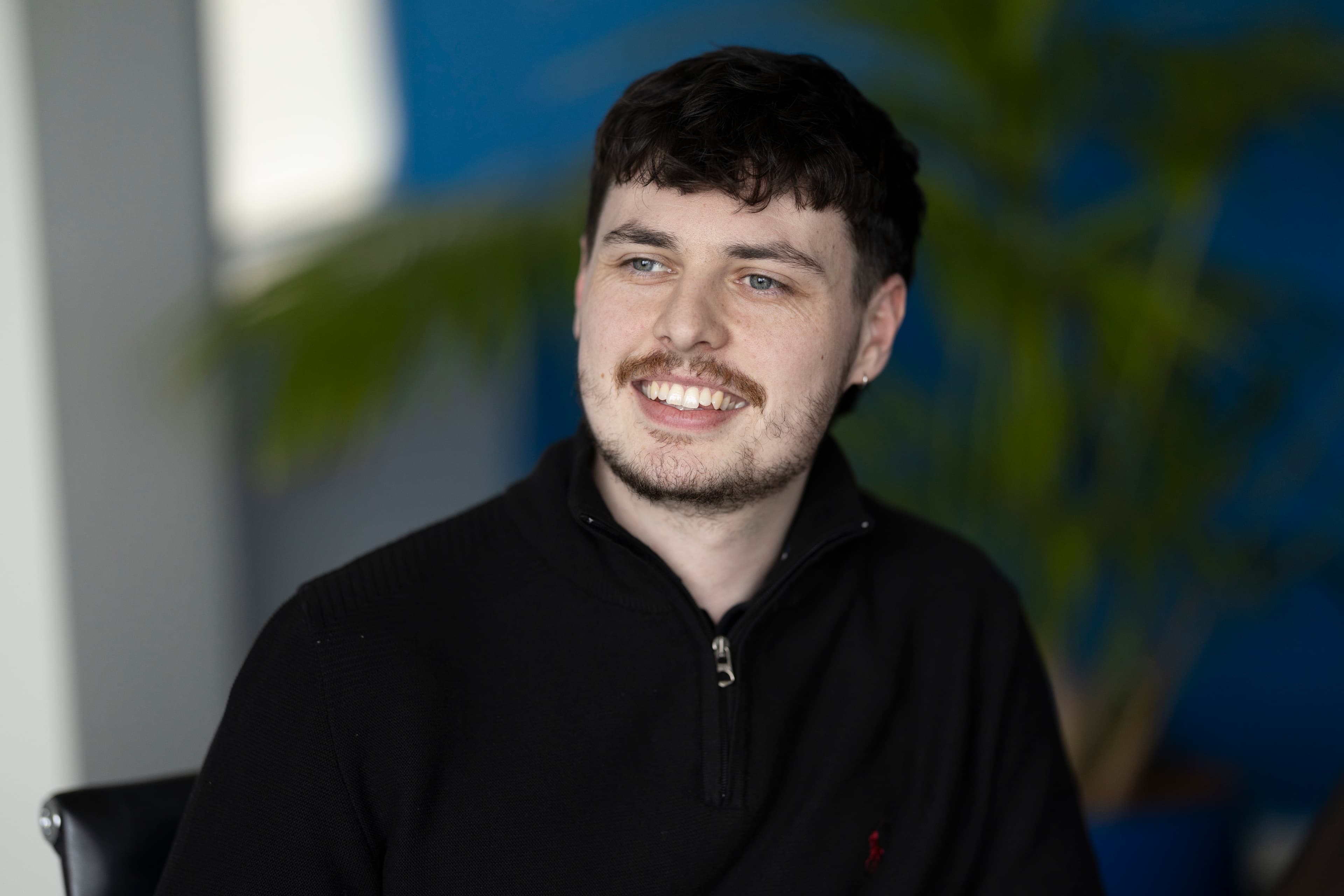 Ean2 — young man with dark curly hair and light beard, dark top, smiling, plant background