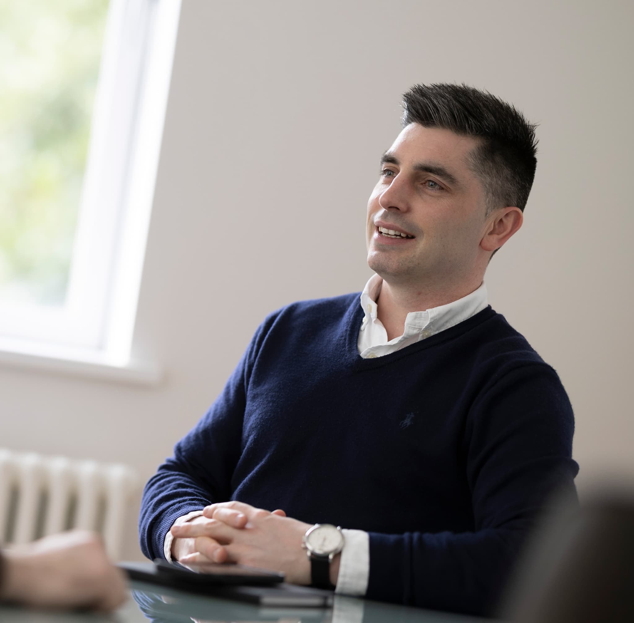 Man with short dark hair, smiling, navy jumper, indoors near a window