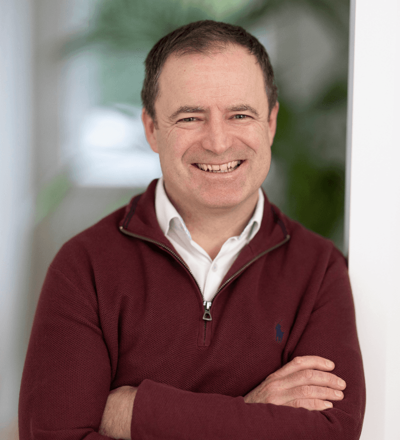 Man in maroon zip jumper, smiling, plant in background