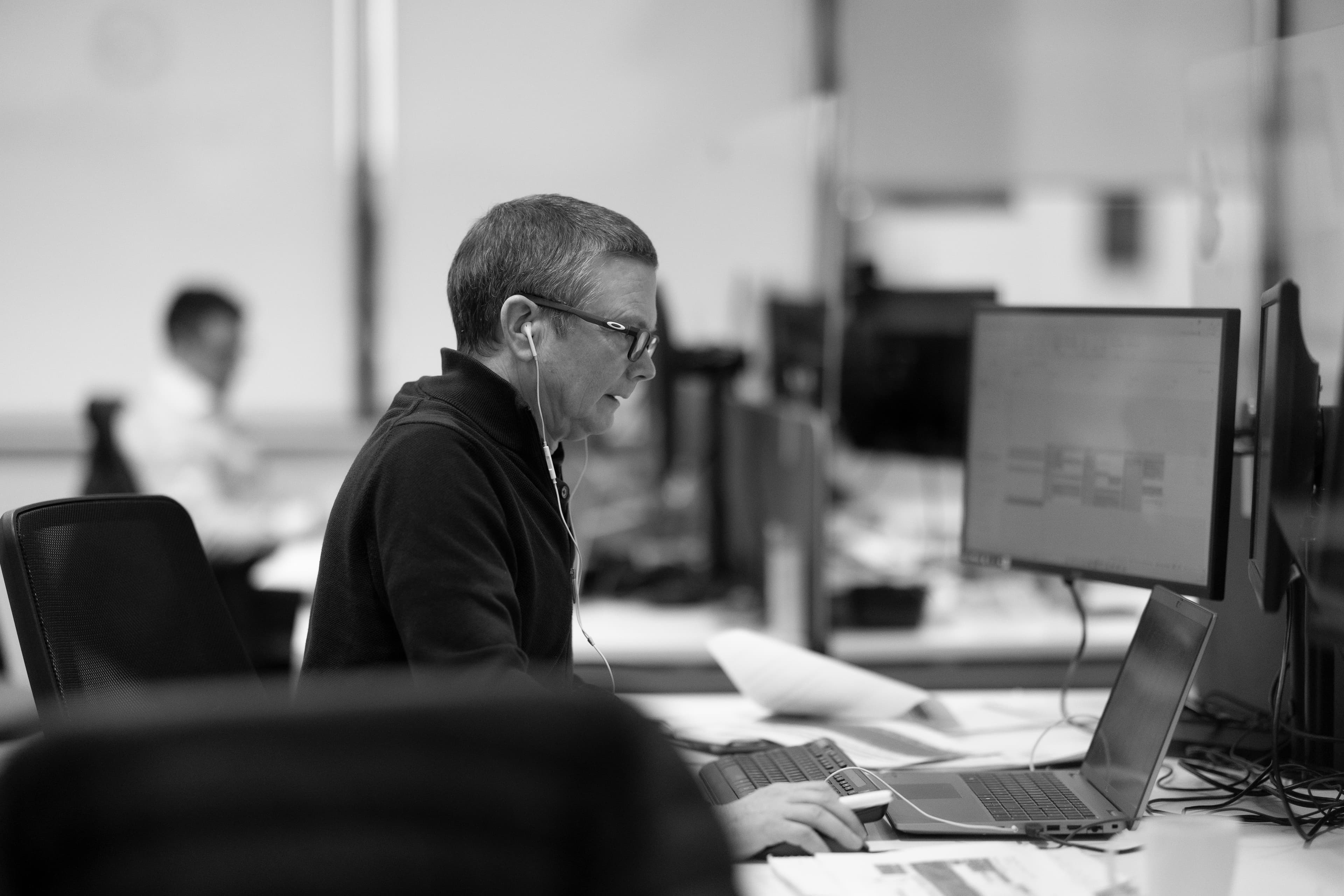 Fergal H — A man with glasses, dark top, working at a desk with monitors, black and white
