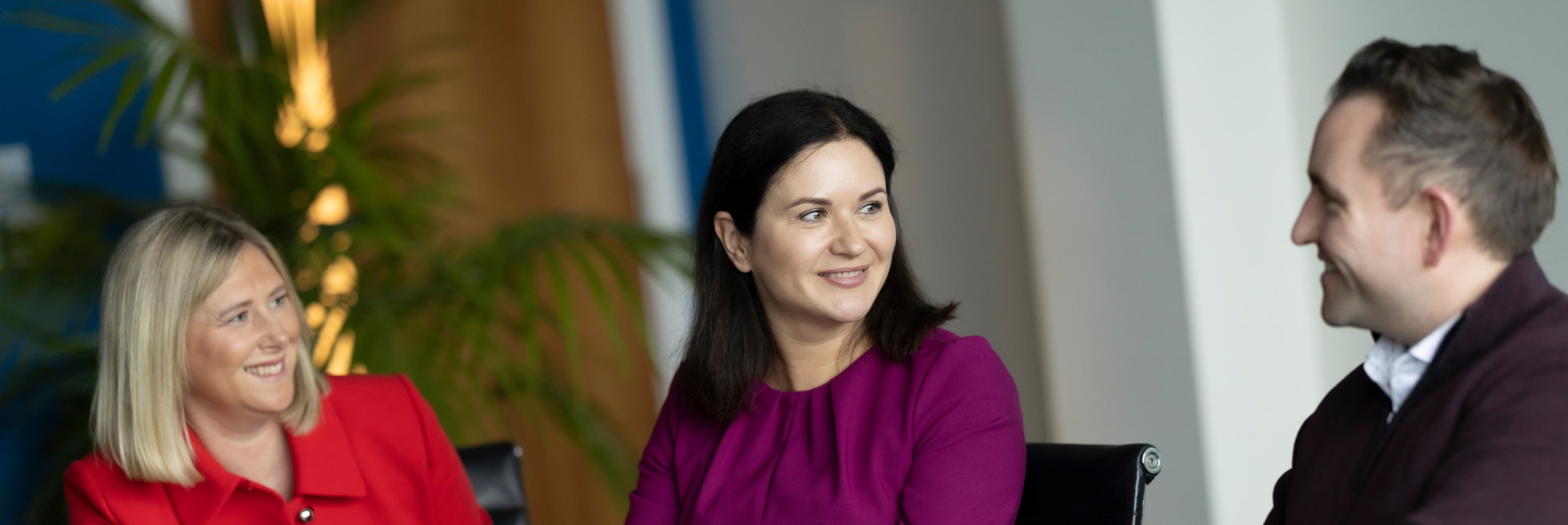 Woman with dark hair, magenta top, smiling, seated indoors near a plant