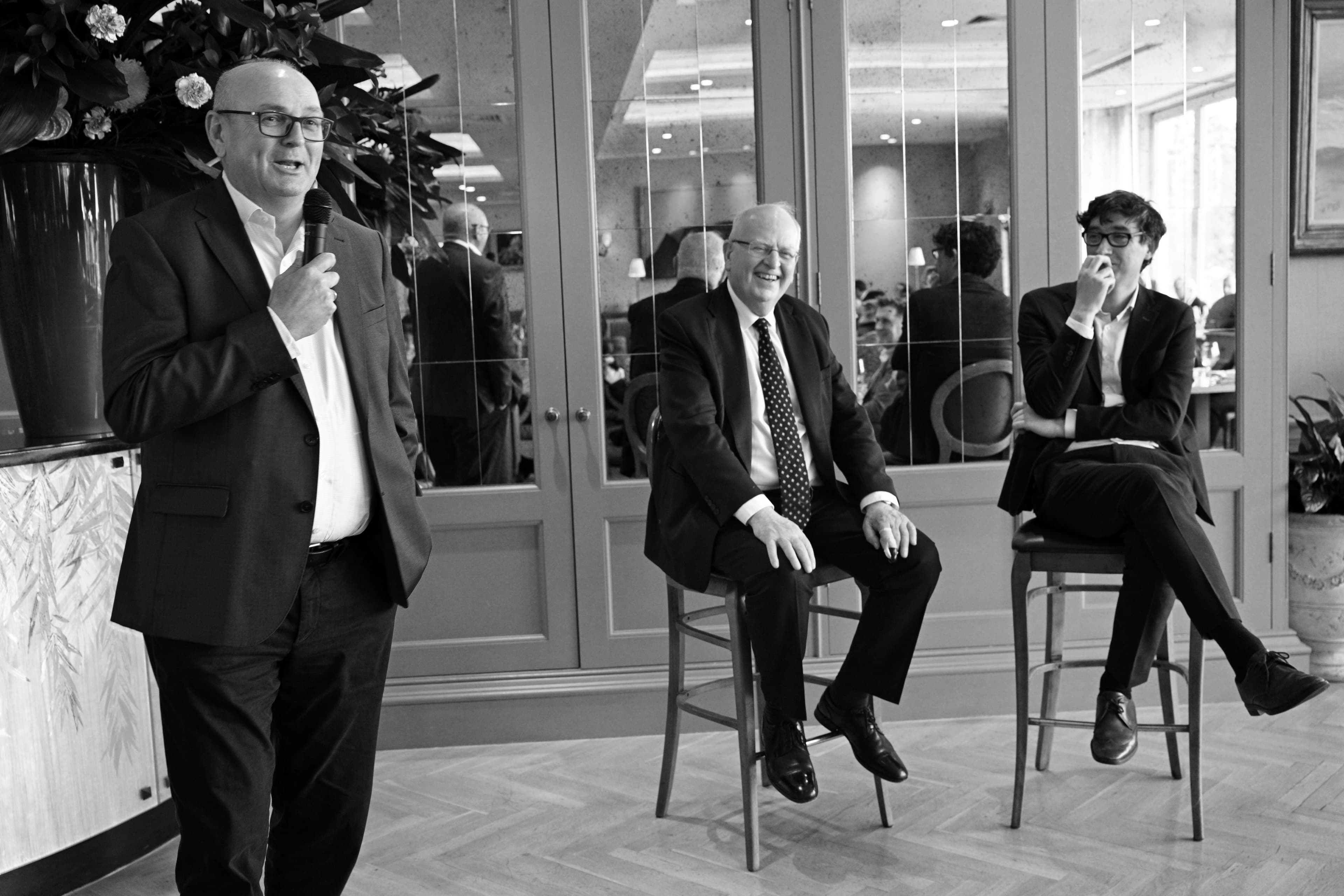 Three men in dark suits in conversation, two seated with a drink, Capitalflow lunch