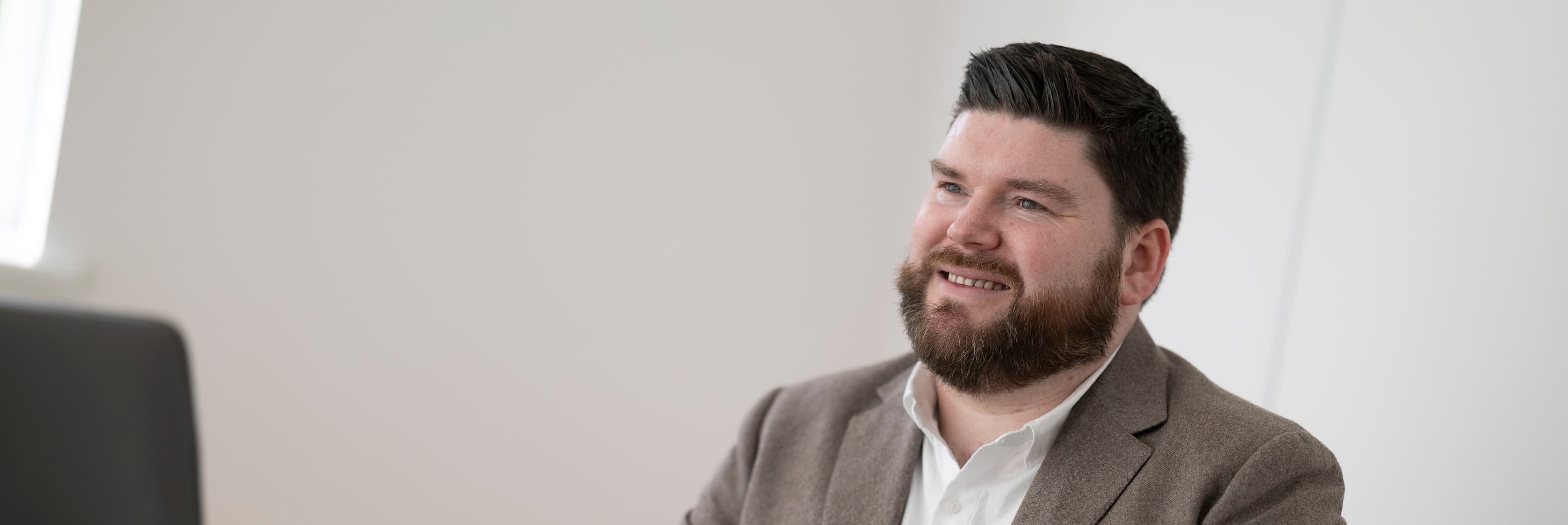 Man with dark beard, smiling, brown jacket, indoors