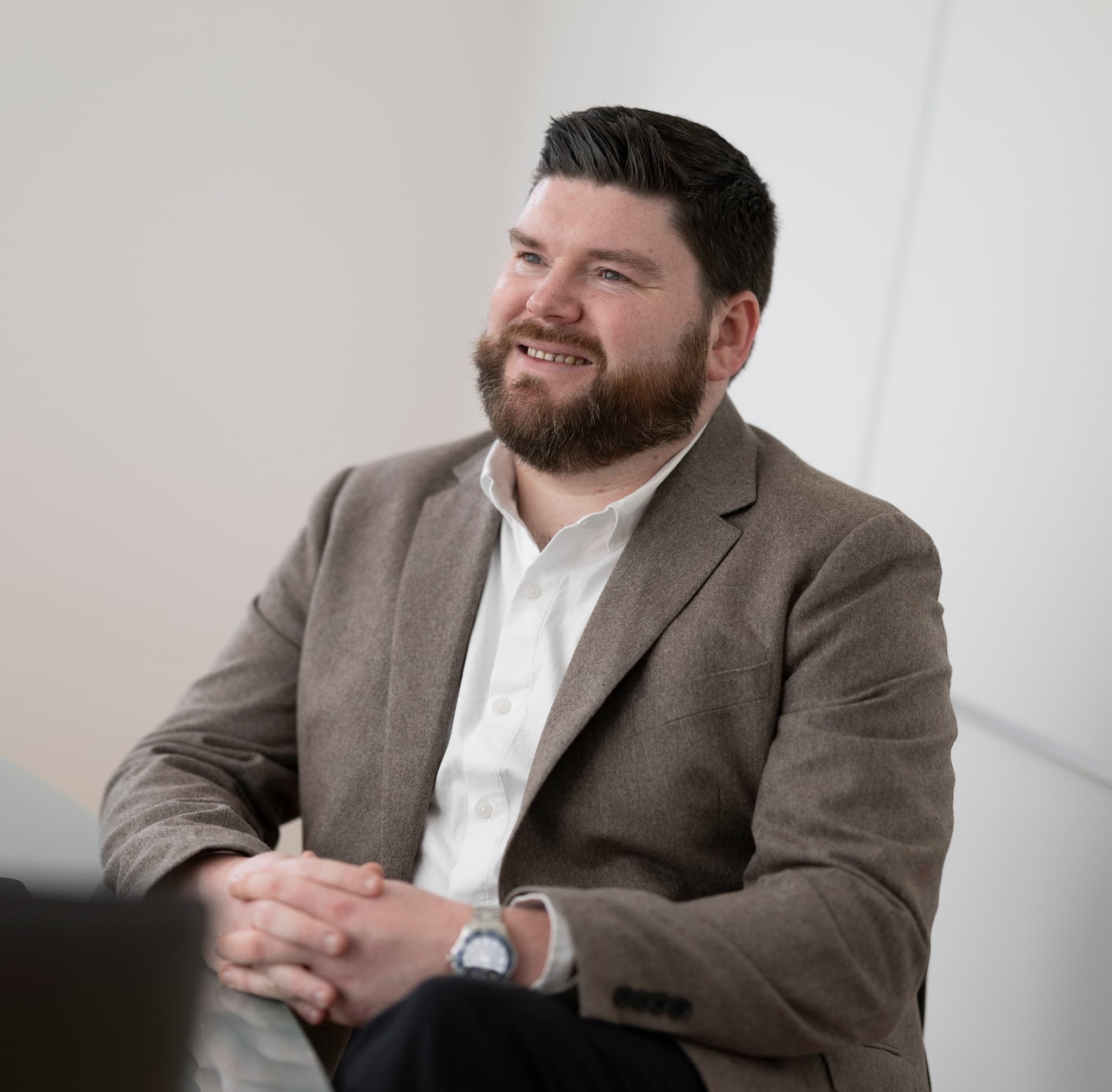 Man with dark beard, smiling, brown jacket, indoors