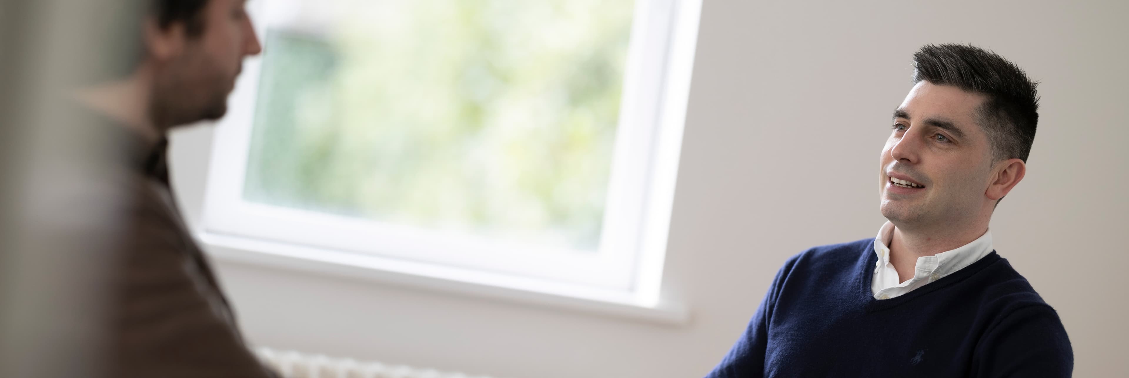 Man with short dark hair, smiling, navy jumper, indoors near a window