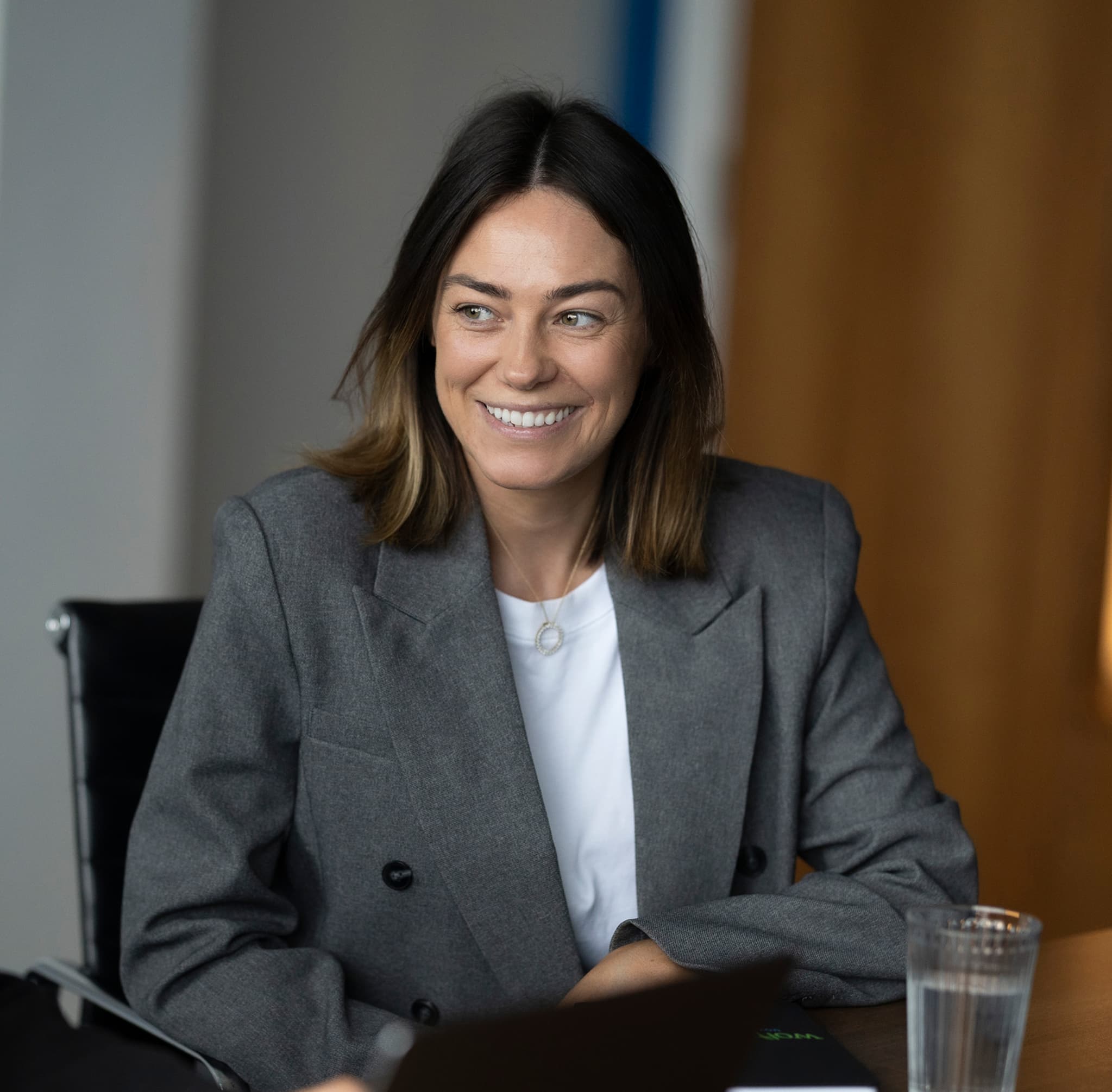 Woman with dark blonde hair, smiling, blazer, indoors, blurred