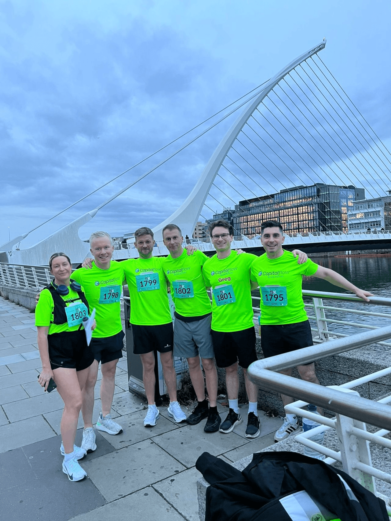 Group in bright green Capitalflow event T-shirts standing on a pedestrian bridge with the Samuel Beckett Bridge cables and Dublin's Grand Canal Dock skyline behind them