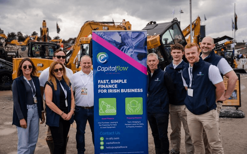 Group of five people (two women in sunglasses, three men) standing beside construction excavators, holding a Capitalflow banner reading "Fast, Simple Finance for Irish Business," overcast sky